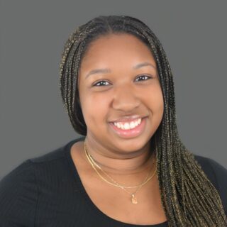 Smiling person with long braided hair wearing a black top and gold necklace against a gray background.