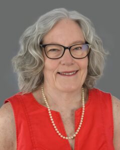 Older woman with gray hair and glasses, wearing a red top and pearls, smiling against a gray background.