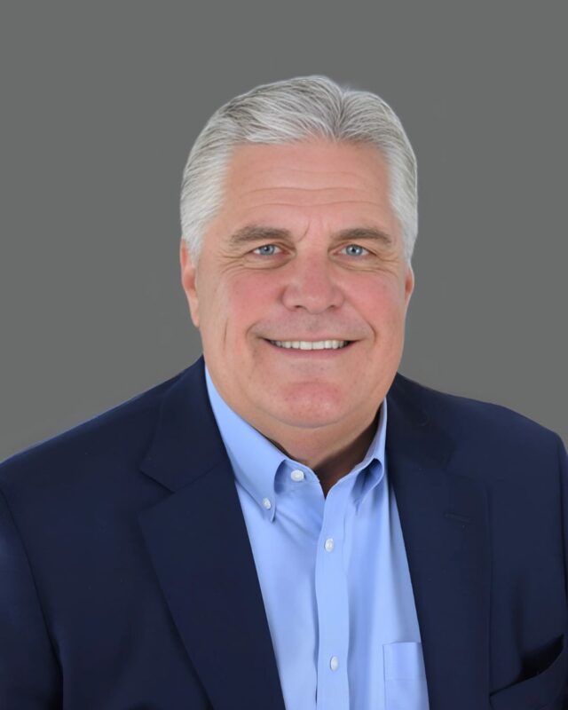 Smiling person with short gray hair wearing a navy blazer and light blue shirt against a neutral background.