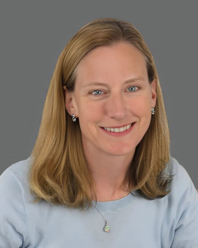 Smiling person with shoulder-length hair wearing a light blue shirt against a gray background.