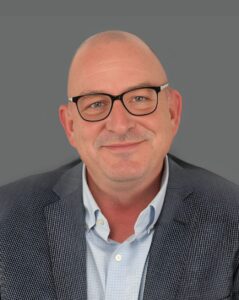 Smiling bald man wearing glasses and a suit jacket against a gray background.