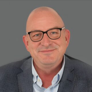 Smiling bald man wearing glasses and a suit jacket against a gray background.