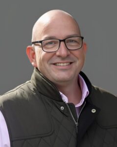 Smiling person wearing glasses and a dark vest against a gray background