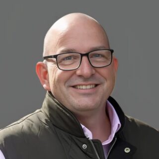 Smiling person wearing glasses and a dark vest against a gray background