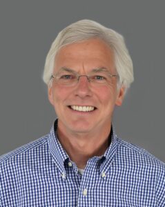 Smiling elderly man with white hair wearing glasses and a blue checkered shirt against a gray background.