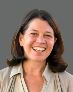 Smiling person with shoulder-length dark hair wearing a beige shirt against a plain gray background.