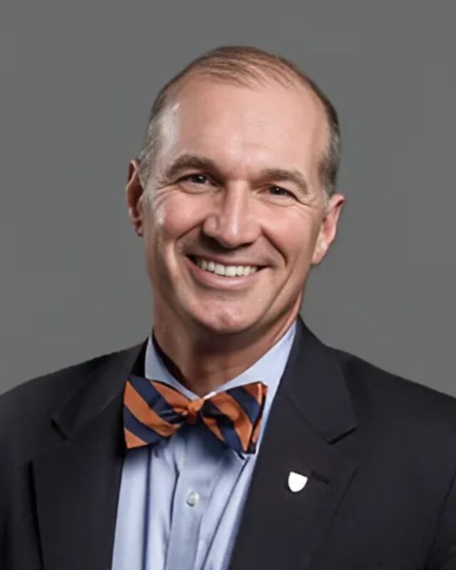 Smiling person wearing a bow tie and a suit against a plain background.