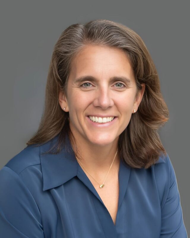 Smiling person with long brown hair wearing a blue shirt against a plain gray background.