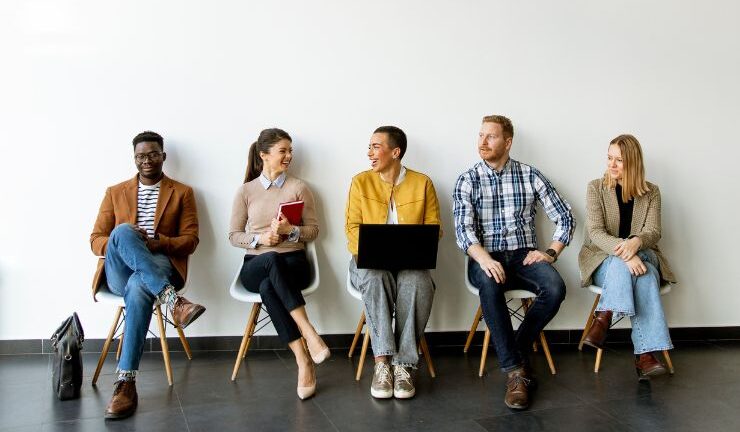 A diverse group of people sitting on chairs against a white wall, engaged in conversation and using electronic devices.