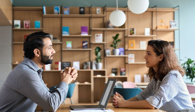 Two people sitting at a desk in an office, having a conversation with a laptop open in front of them.
