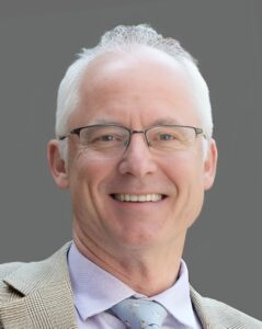 Portrait of a smiling man wearing glasses and a light-colored suit against a gray background.