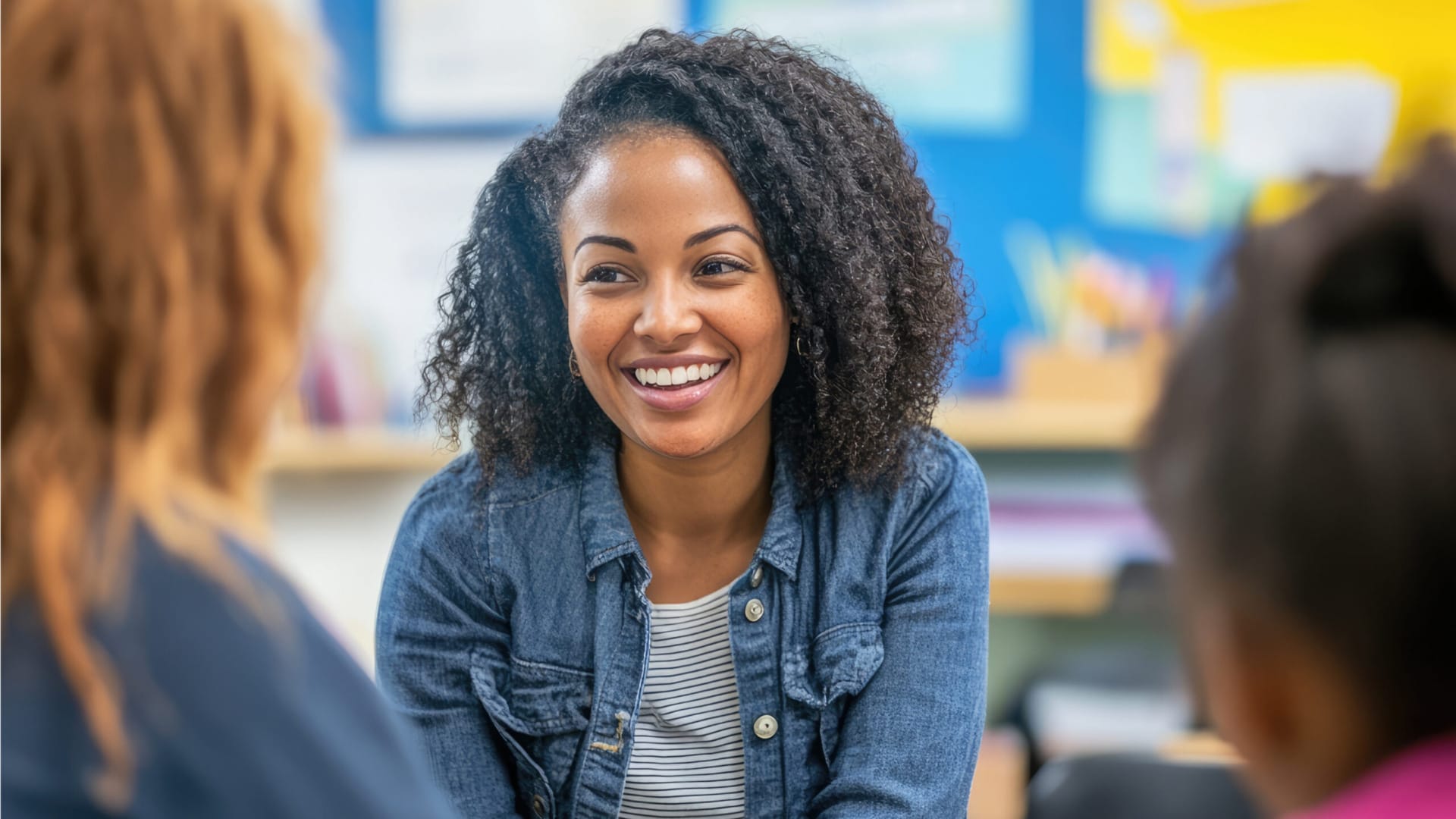 Smiling woman with curly hair sitting at a table in a classroom, engaging in conversation with blurred figures nearby.
