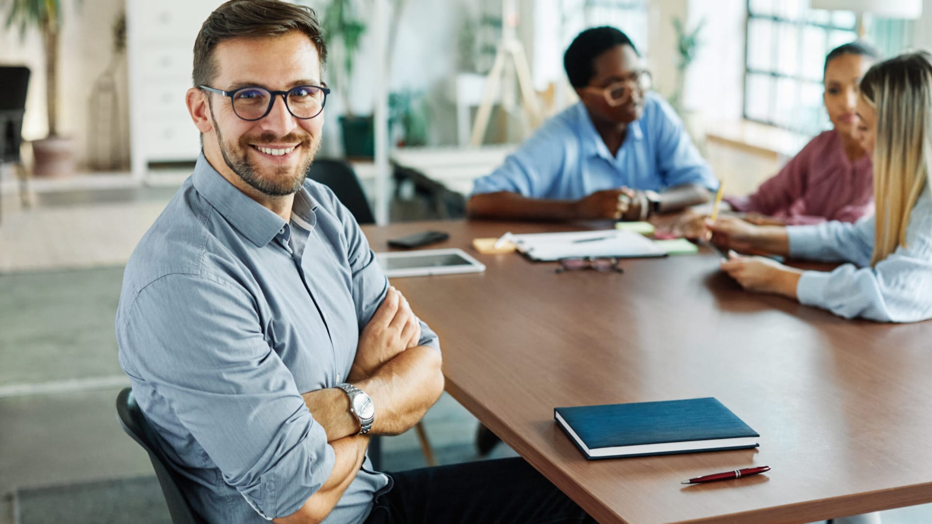 Man smiling while sitting at a conference table with colleagues working in the background.