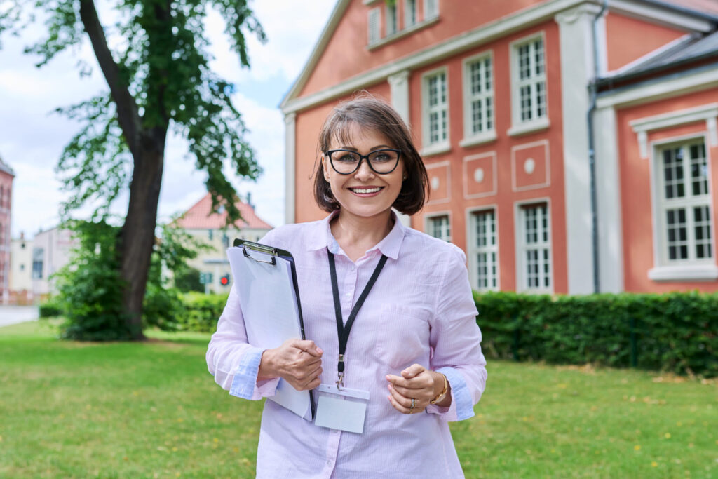 Smiling woman with short hair holding a clipboard, standing outside a red brick building with green trees in the background.
