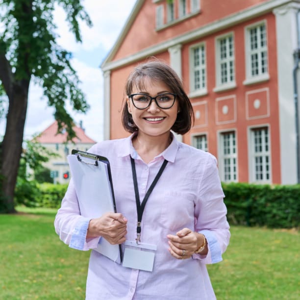 Smiling woman with short hair holding a clipboard, standing outside a red brick building with green trees in the background.