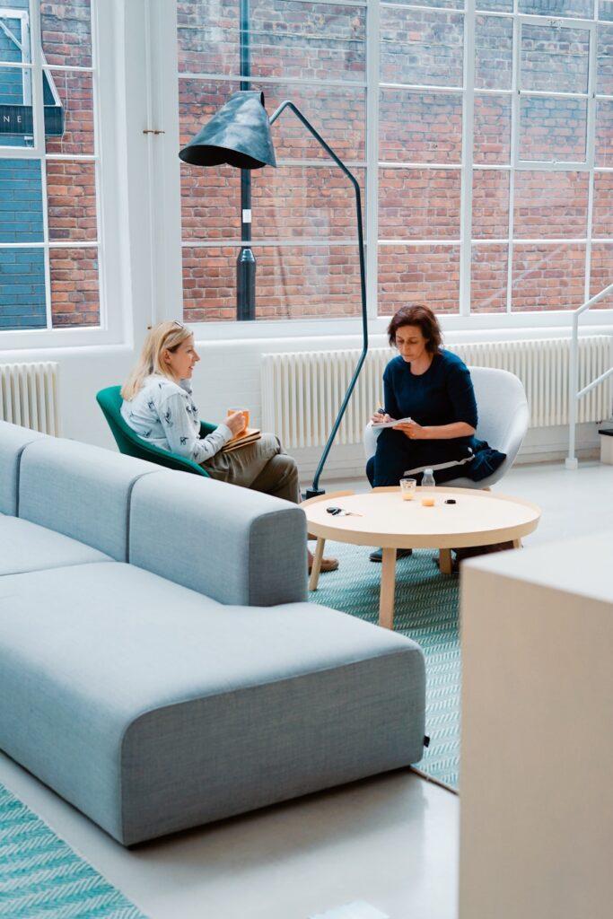 Two people sitting in a modern office space, engaged in conversation near a round table with a couch in the foreground.