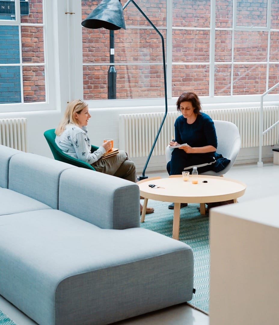 Two people sitting in a modern office space, engaged in conversation near a round table with a couch in the foreground.