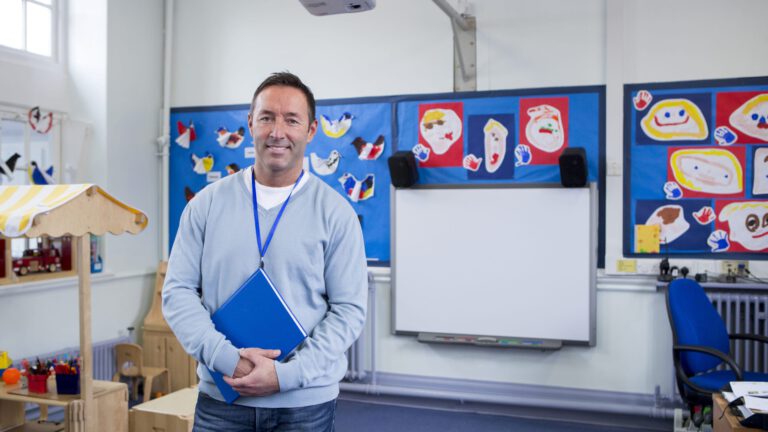 A teacher standing in a classroom in front of a whiteboard, holding a blue folder and wearing a lanyard, with decorations behind.