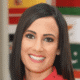 Smiling woman with long dark hair in a red shirt, standing in front of international flags.