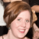 Smiling person with short hair at an event, wearing a dress, surrounded by other people in the background.
