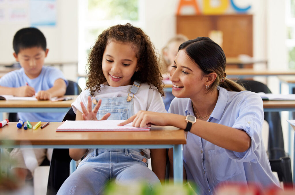 Teacher helping a young girl with curly hair at her desk in a classroom, both smiling and engaged in a learning activity.