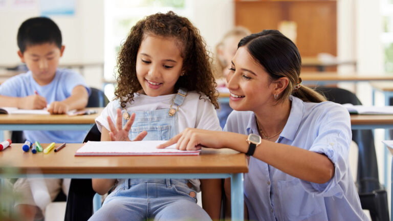 Teacher helping a young girl with curly hair at her desk in a classroom, both smiling and engaged in a learning activity.