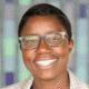 Smiling person wearing glasses and a plaid blazer against a blurred, colorful background