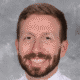 Smiling man with a beard and short hair wearing a white shirt against a gray background.