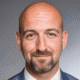 Smiling man with a bald head and a beard, wearing a suit and tie, in front of a neutral background.