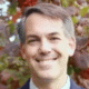 Smiling person standing outdoors with blurred autumn leaves in the background.