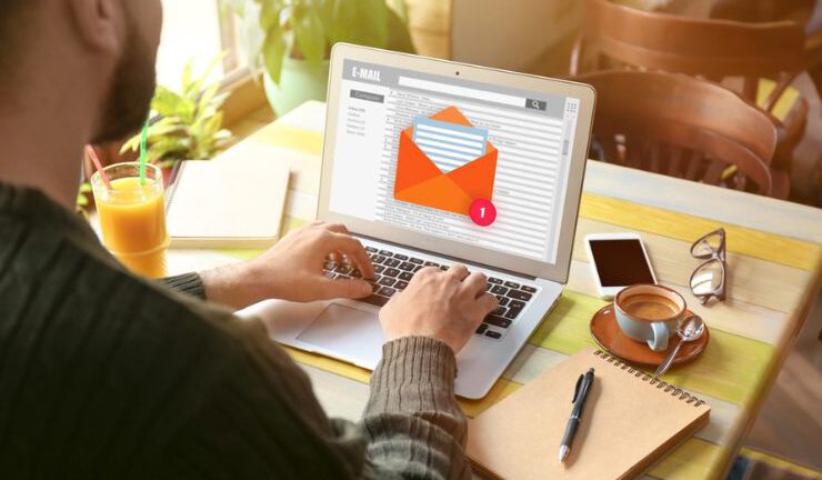 Person typing on a laptop with an email notification on screen, surrounded by a coffee cup and office supplies on a desk.