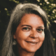 Smiling woman with long gray hair and earrings, outdoors with blurred greenery in the background.