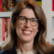 Smiling person with glasses and long hair in front of a bookshelf background.