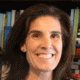 Smiling person with brown hair, wearing a dark shirt, standing in front of a bookshelf filled with colorful books.