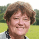 Smiling person standing outside with a blurred green field and trees in the background.