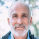 Older man with a beard smiling outdoors with blurred trees in the background.