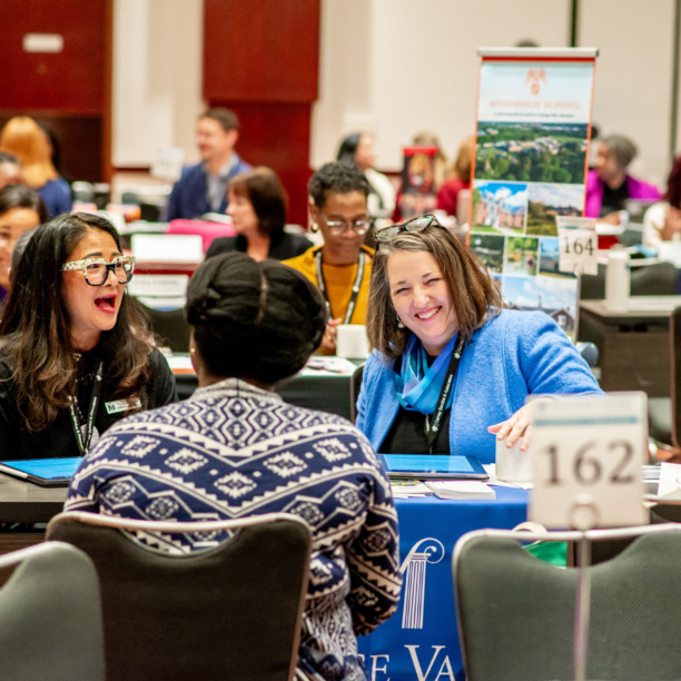 People at a conference table engaged in conversation with brochures and signs on the table in a busy conference room.