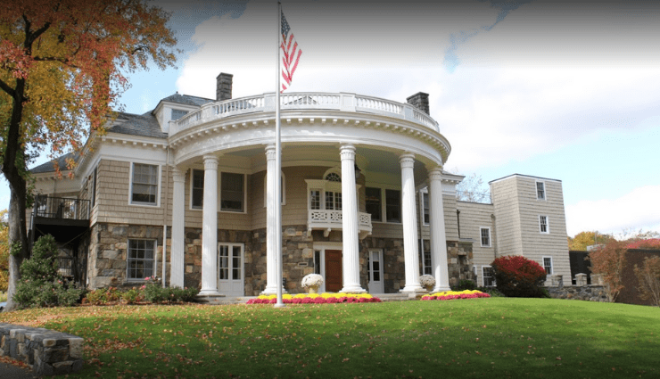Large historic building with round columns and an American flag in front, surrounded by a lawn and trees.