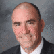 Man in a suit smiling, bald, with a gray background.