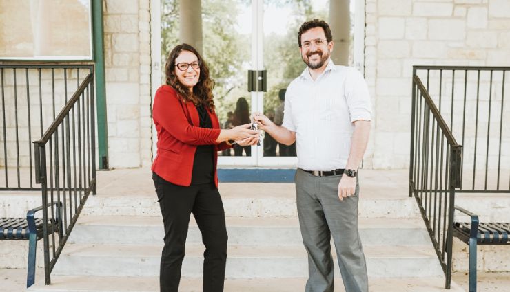 Two people standing on steps, shaking hands and exchanging a key in front of a building entrance.