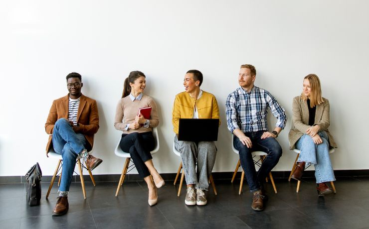 Diverse group of five people sitting on chairs, chatting and using devices against a white wall.