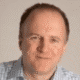Smiling person in a checkered shirt with a neutral background.