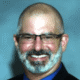 Man with glasses and a beard smiling, wearing a suit and tie against a gray background.