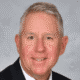 Smiling man in a suit and tie with gray hair against a gray background.