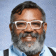 Smiling person with glasses and a beard, wearing a light blue shirt against a plain background.