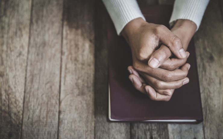 Person with clasped hands resting on a book on a wooden table