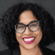 Woman with curly hair and glasses, smiling, wearing bright lipstick against a dark background.