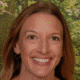 Smiling person with long hair standing in front of a green leafy background.
