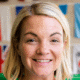 Smiling person with short blonde hair, wearing green top, background with colorful flags.