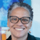 Smiling person with short curly hair and glasses, wearing earrings, in front of a colorful abstract background.